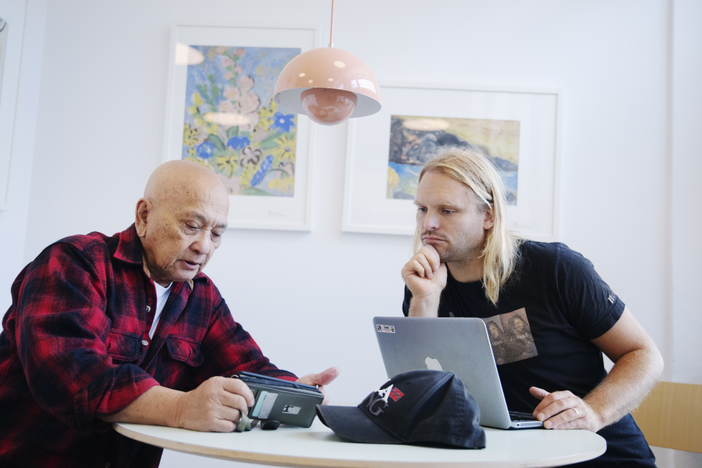 Ett foto av en senior person sitter tillsammans med en yngre person framför mobiltelefon och dator vid ett bord.